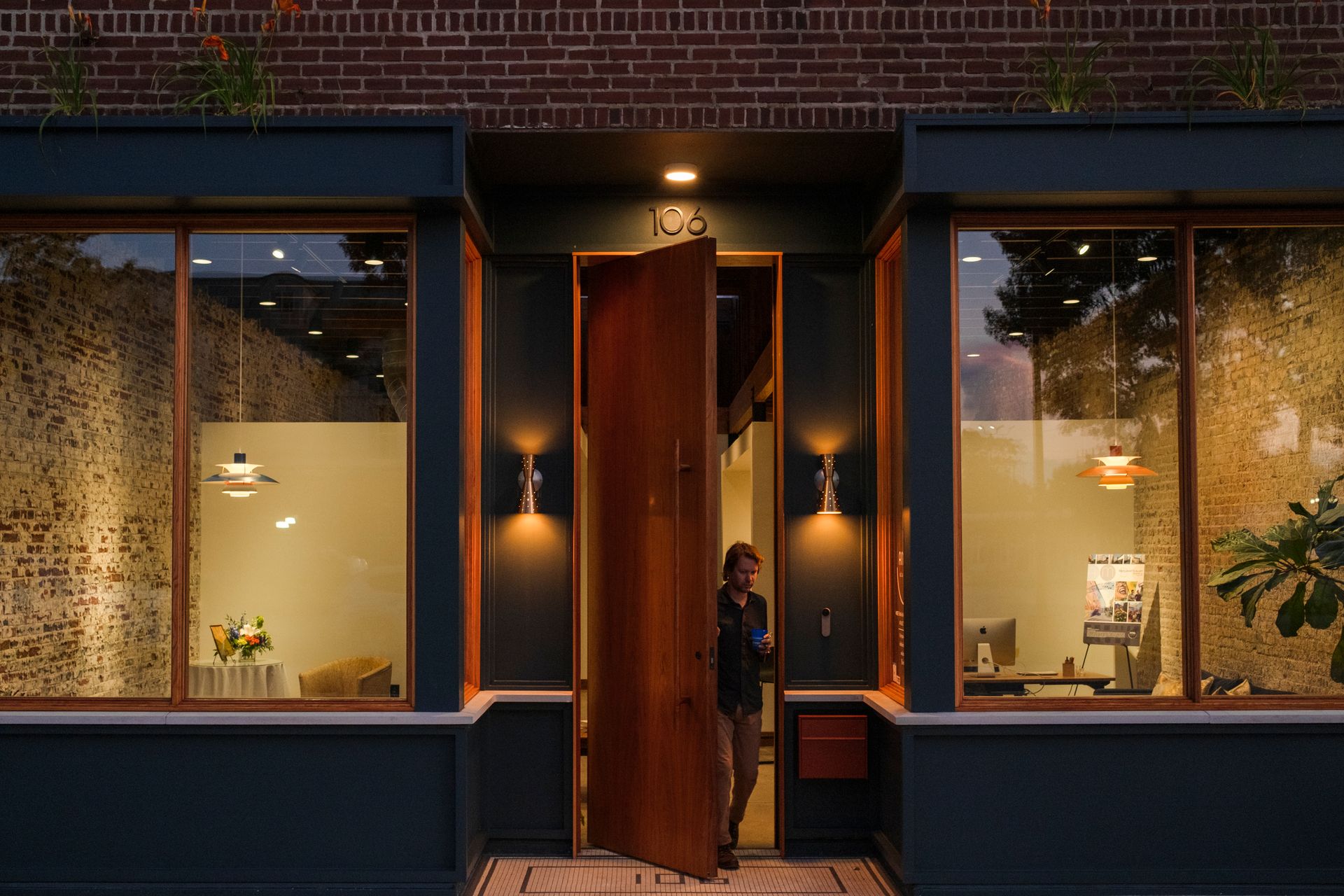 Exterior of a building with an open wooden door. A person stands in the doorway. Flanking windows on either side. Dark green trim, brick facade.