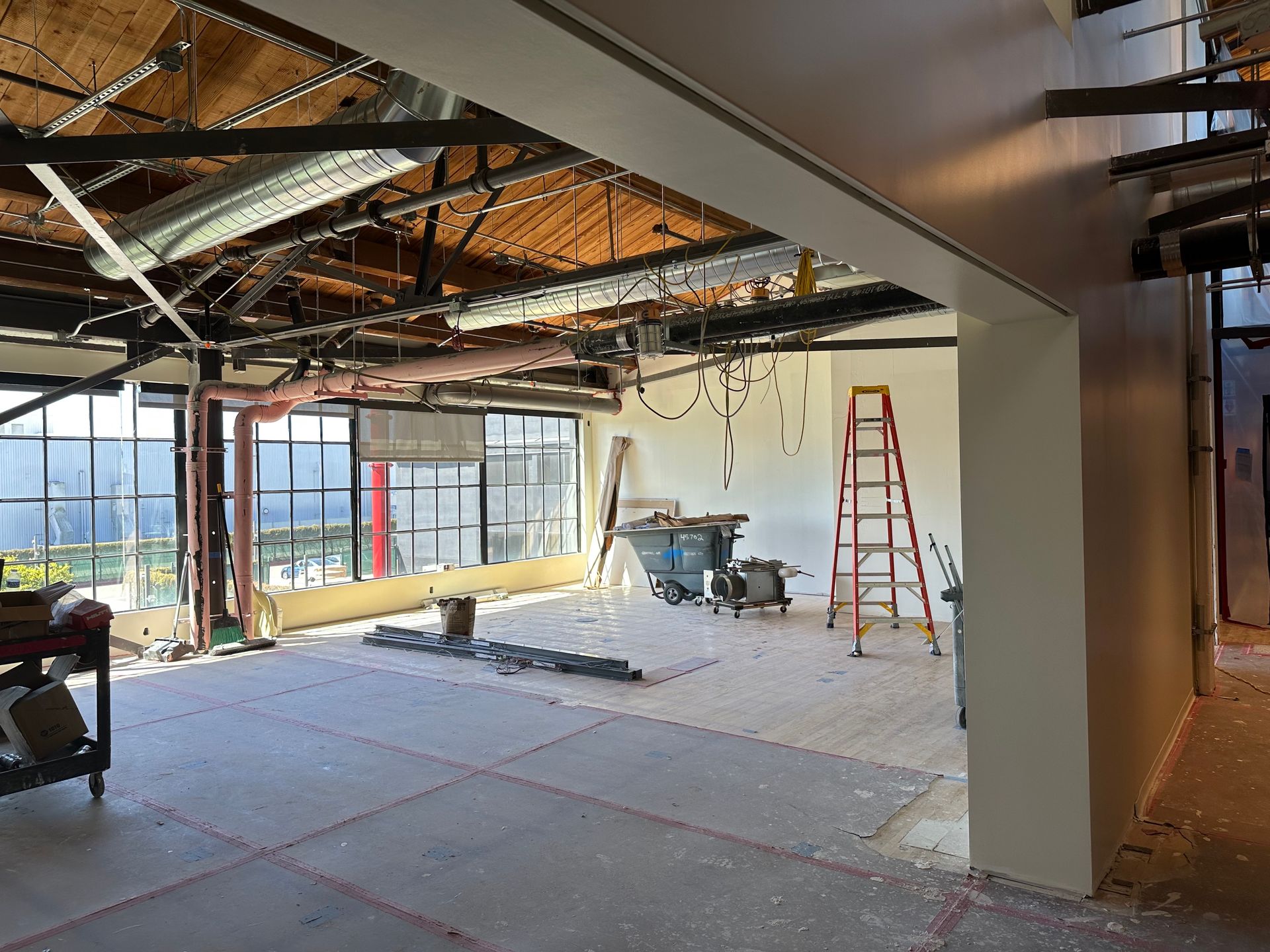 Interior under construction: exposed ceiling, tools, ladder, large windows, light-colored walls, and debris on the floor.
