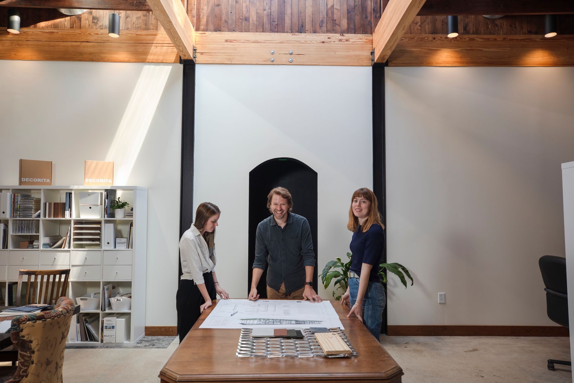 Three people review blueprints at a table in a sunlit office.
