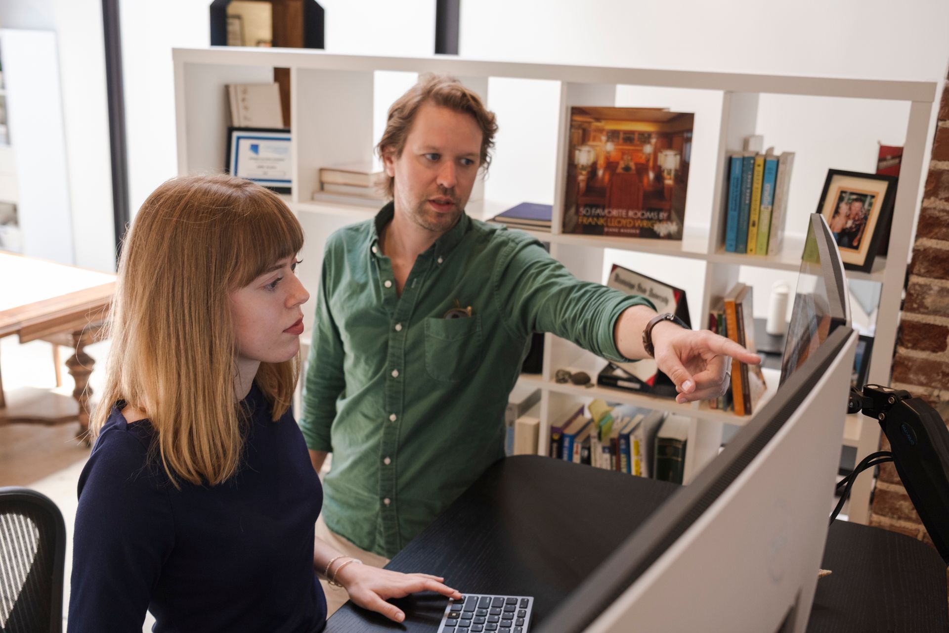 Man points at computer screen, showing woman. Office setting with bookshelves.