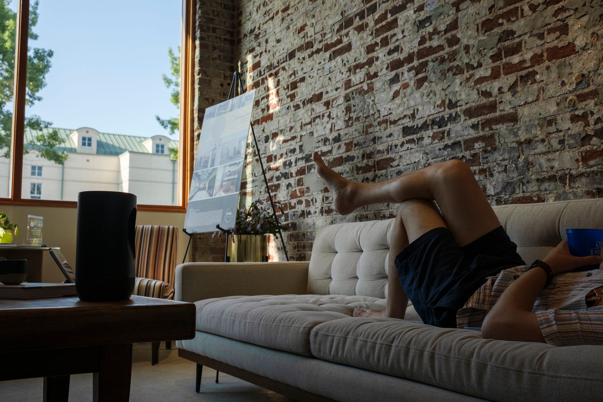 Man relaxing on couch in a loft apartment, legs up, holding a cup, in front of a brick wall.