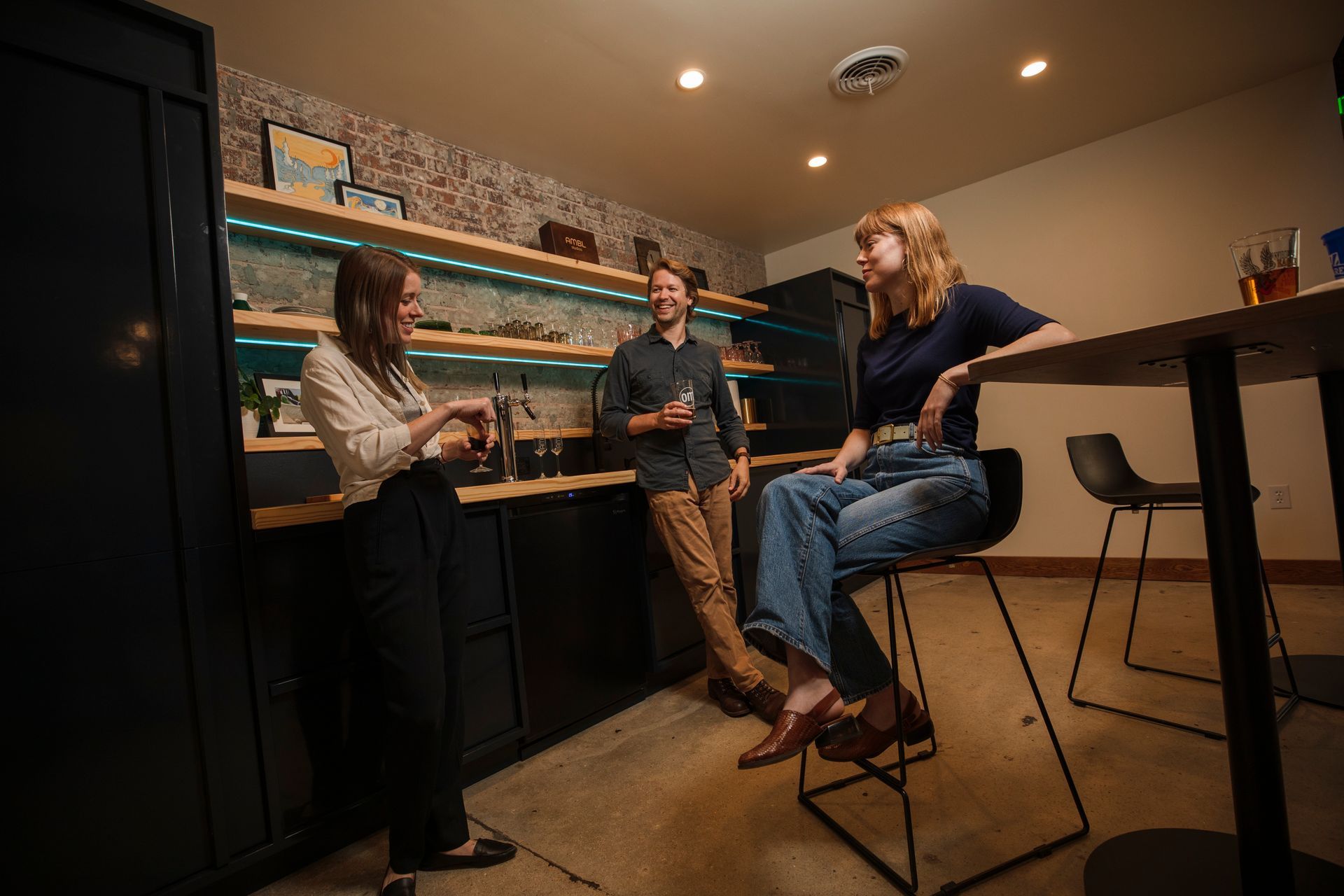 Three people in a small bar area, one seated, two standing, socializing and smiling. Brick wall and wooden shelves behind them.
