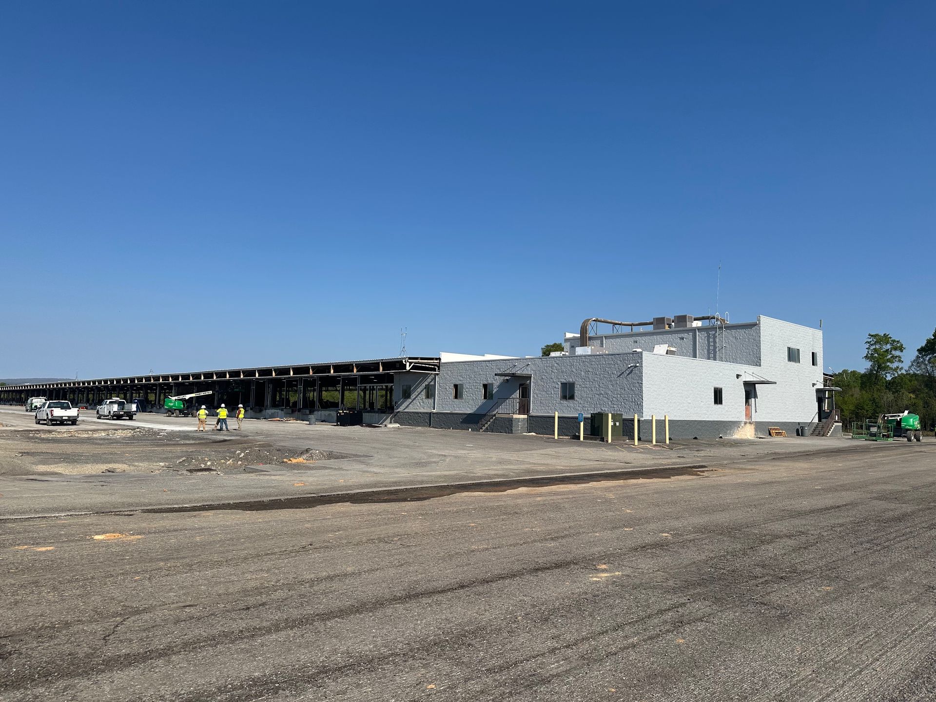 Wide dirt lot with a long low building and white structures under a clear blue sky.