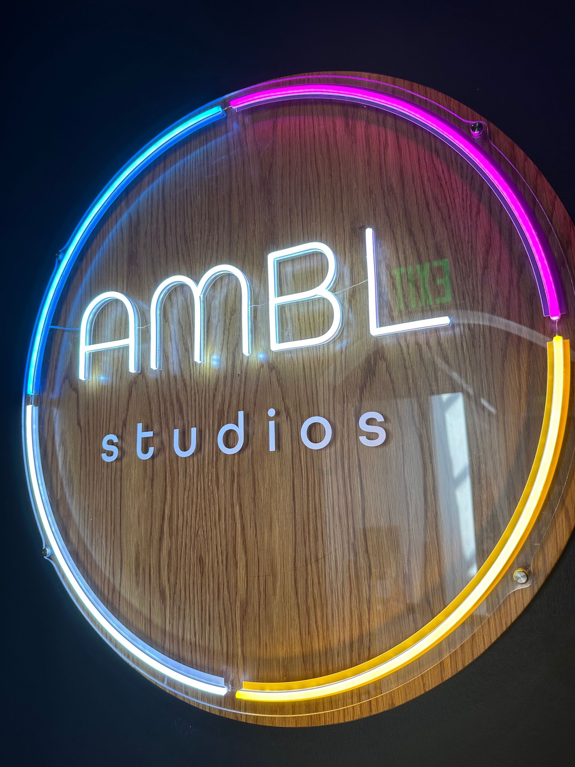 Circular AMBL studios sign with neon multi-colored outline, on wooden background.