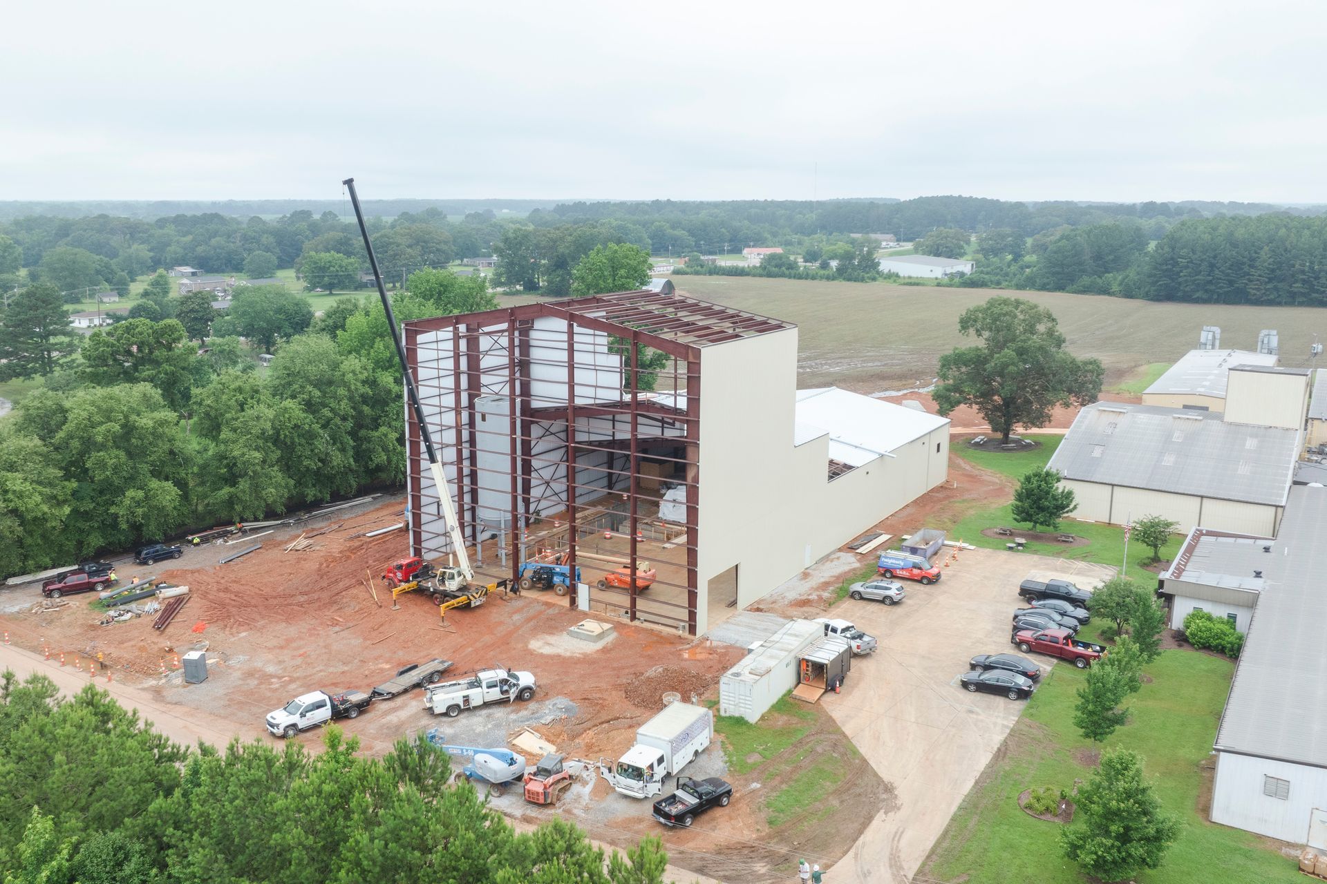 Construction site with steel frame building, crane, and vehicles on dirt, surrounded by trees and fields.