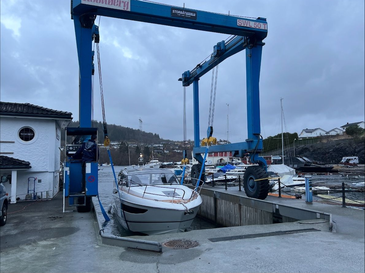 En blå båtheis som løfter en hvit båt i en havn, med grønne åser i bakgrunnen.