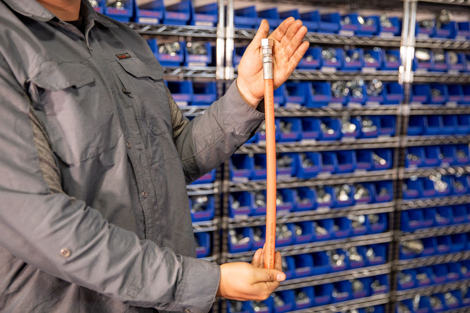 Person in gray shirt holding a long, copper-colored hose with metal fittings, in front of shelves of blue bins.