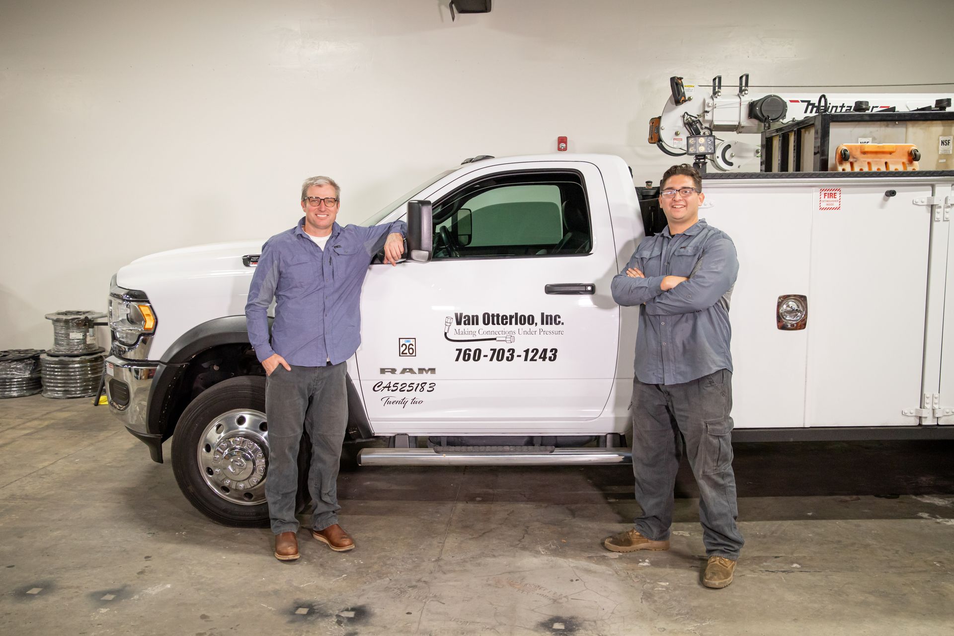 Two men stand by a white work truck with company logo; one leans, the other arms crossed.