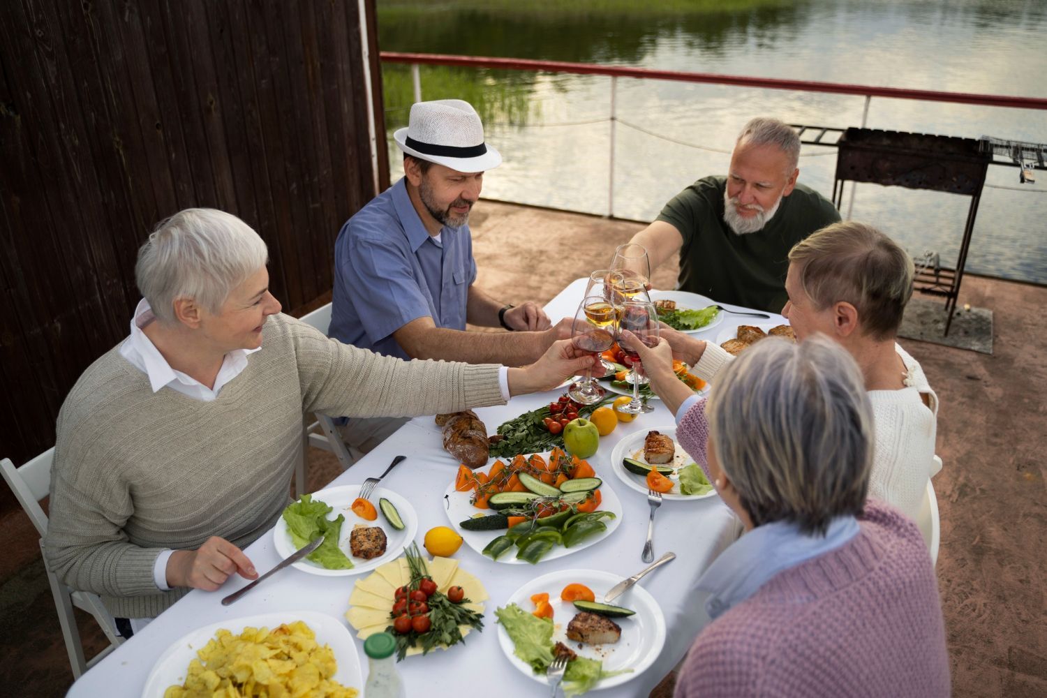 Five people share a meal outdoors at a long table with dishes, flowers, and drinks
