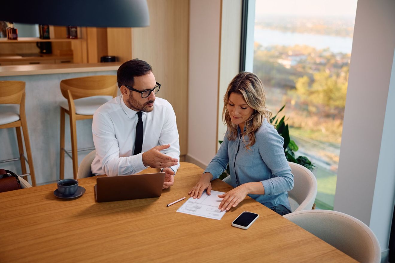 Two coworkers reviewing documents at a wooden table with a laptop and smartphone in a bright office