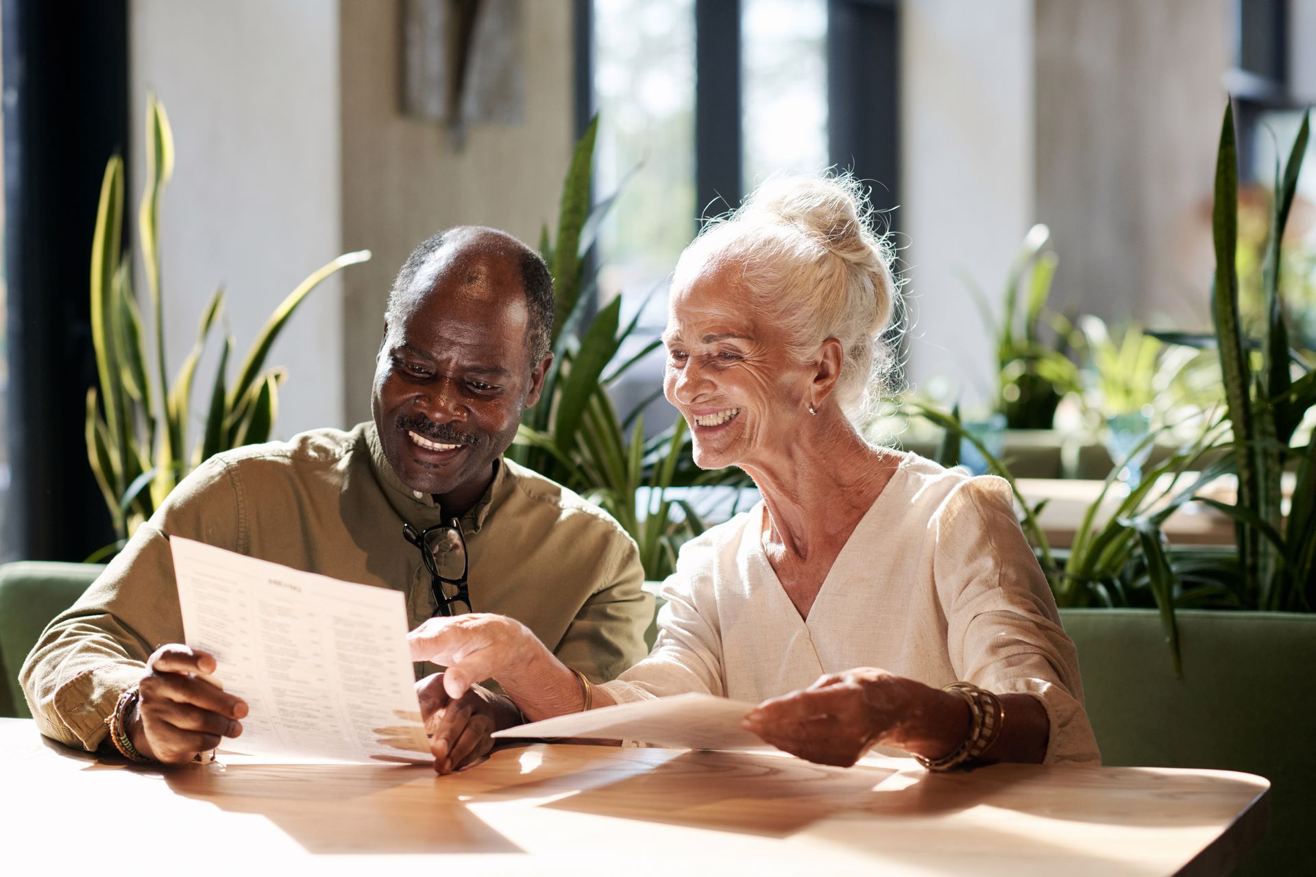 Two people smiling and reading a document together at a sunlit table with plants nearby