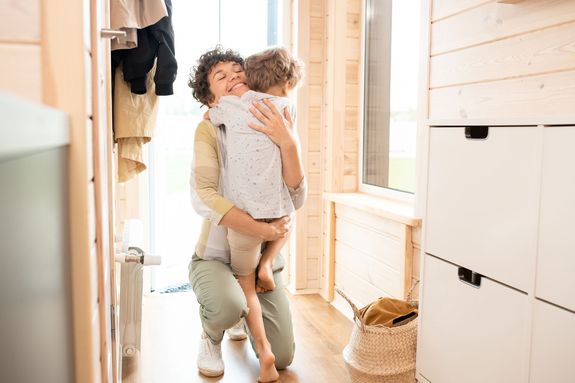 Person holding a child in a bright hallway, standing near open windows and white cabinets.
