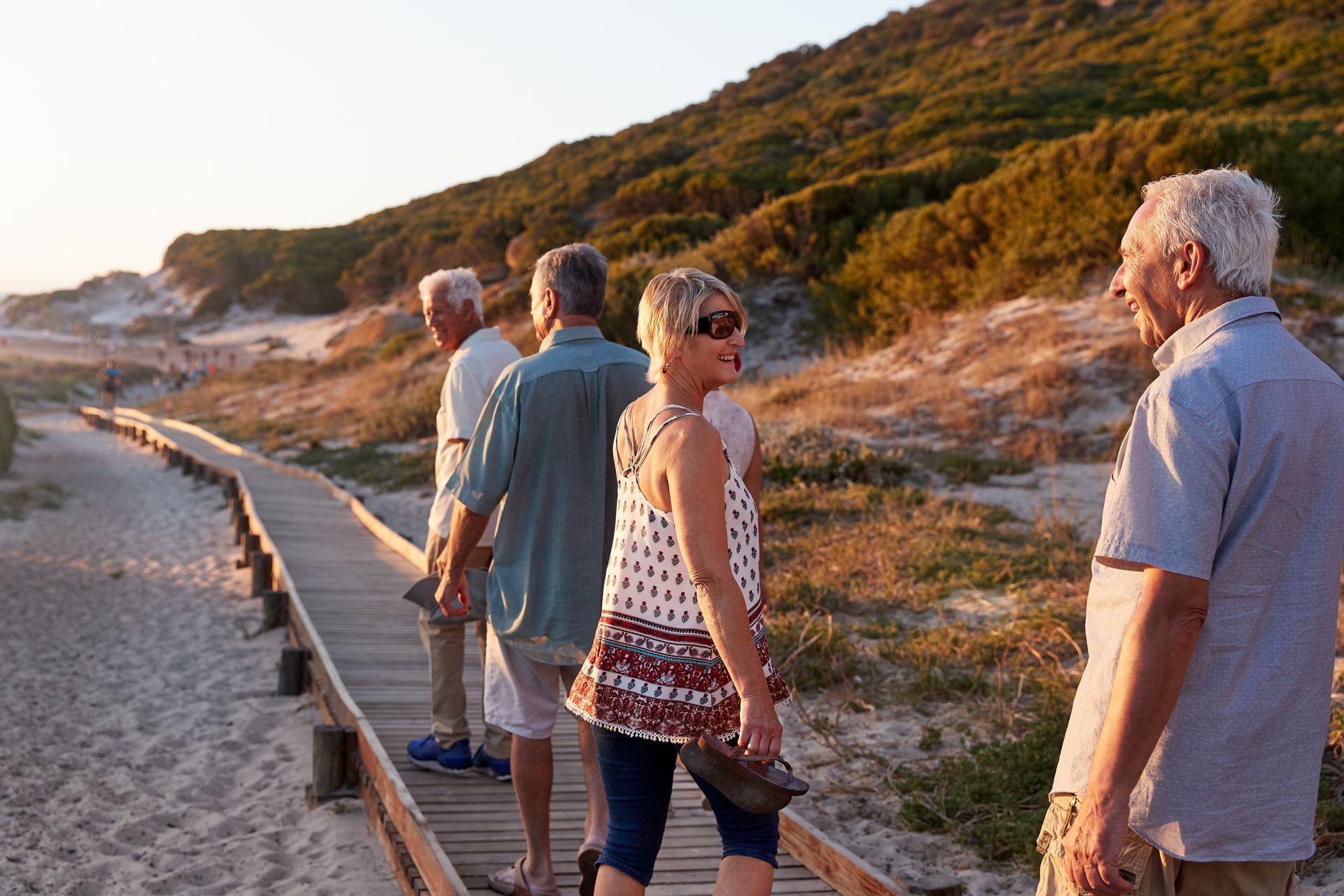 Four people walking along a paved coastal path beside dunes at sunset