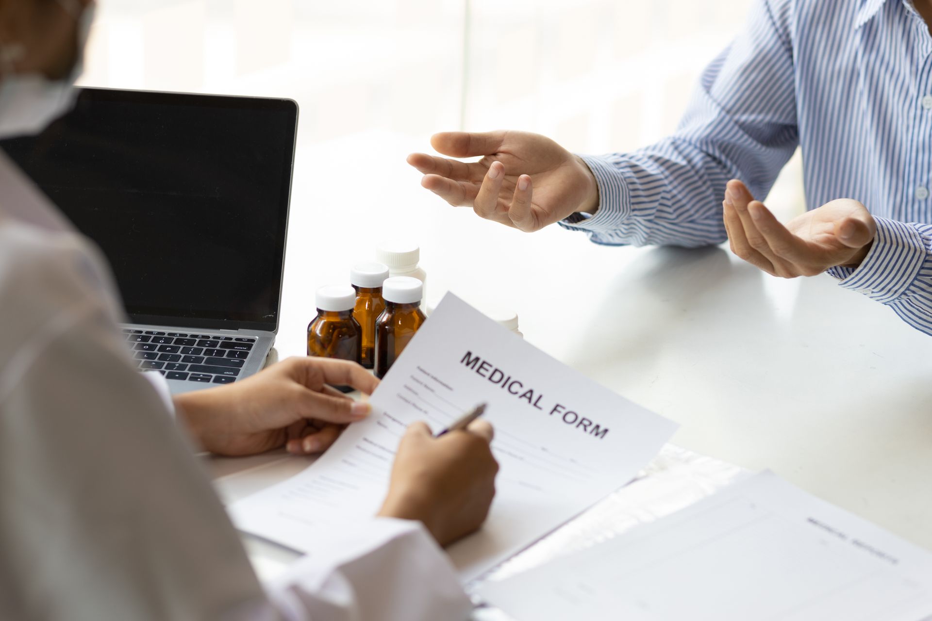 People discussing a medical form at a desk with pill bottles and a laptop nearby