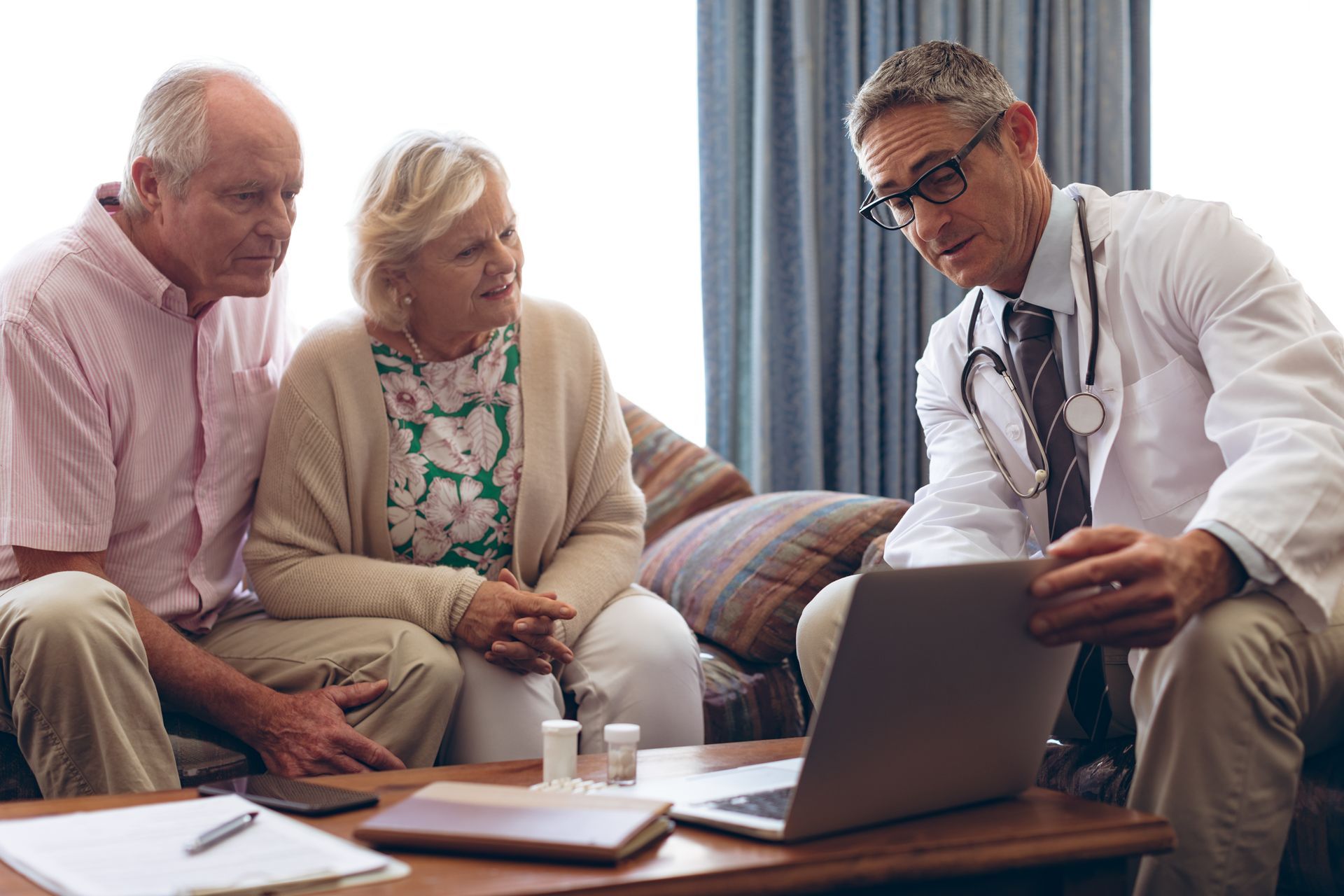Doctor showing laptop to two seated patients in a living room, with papers and coffee table nearby