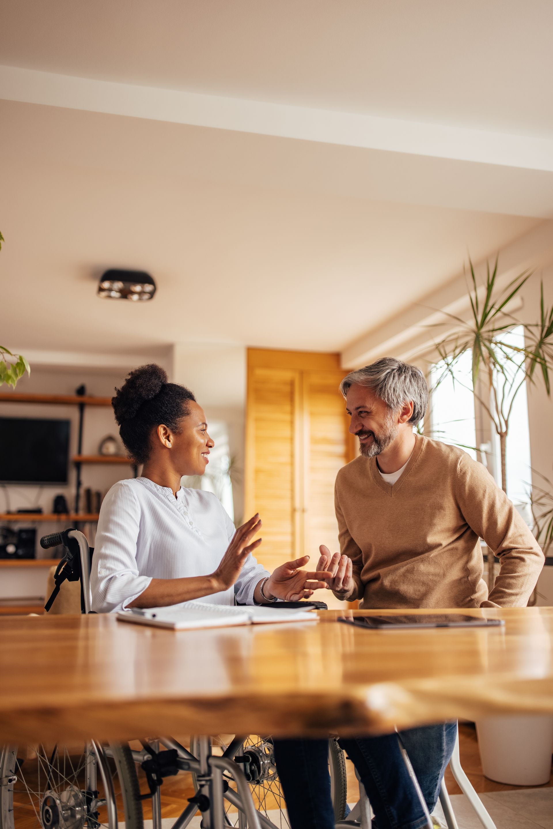 Two people talking at a wooden table in a bright living room with plants and sunlight