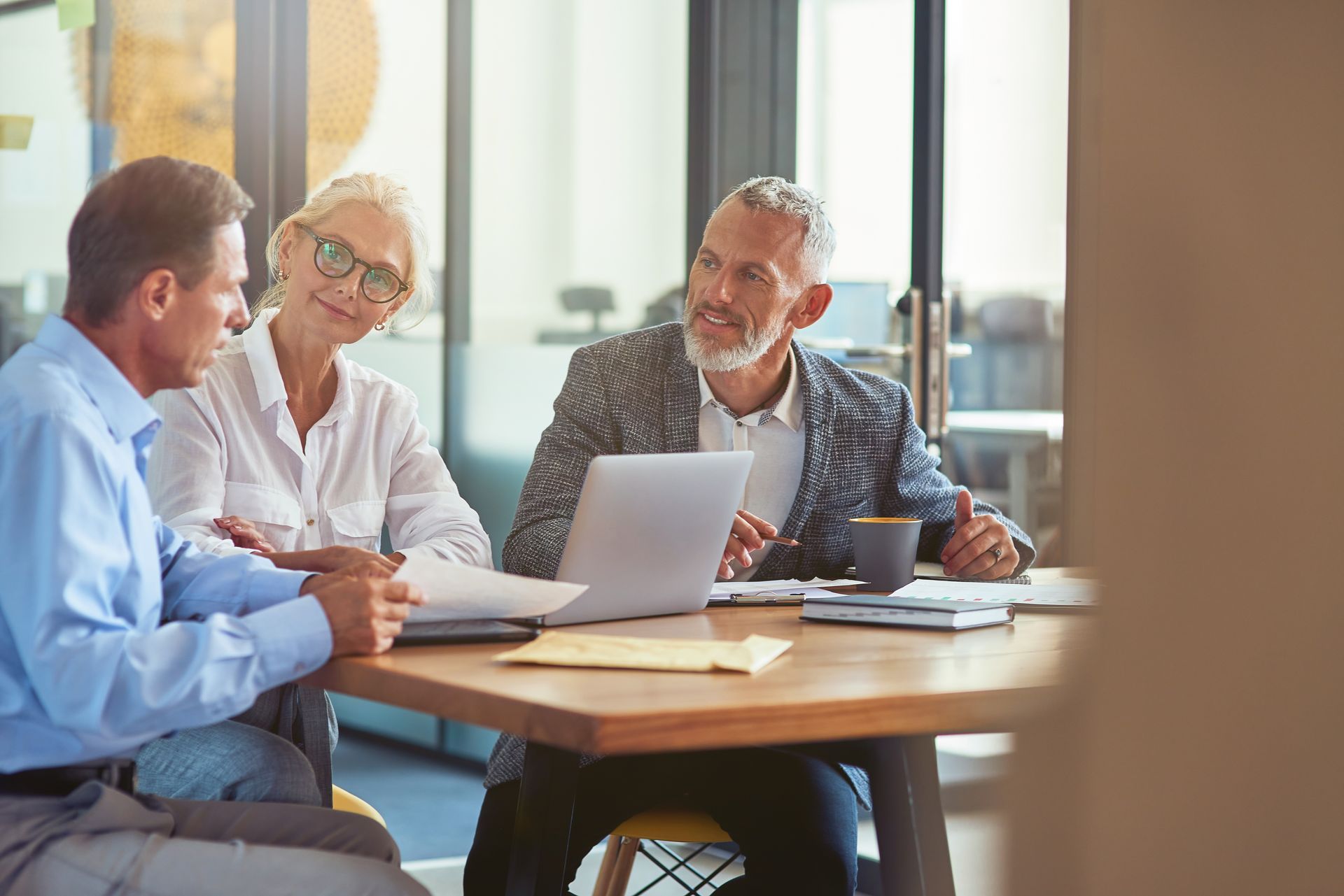 Three people in a meeting around a laptop at a wooden table in a bright office