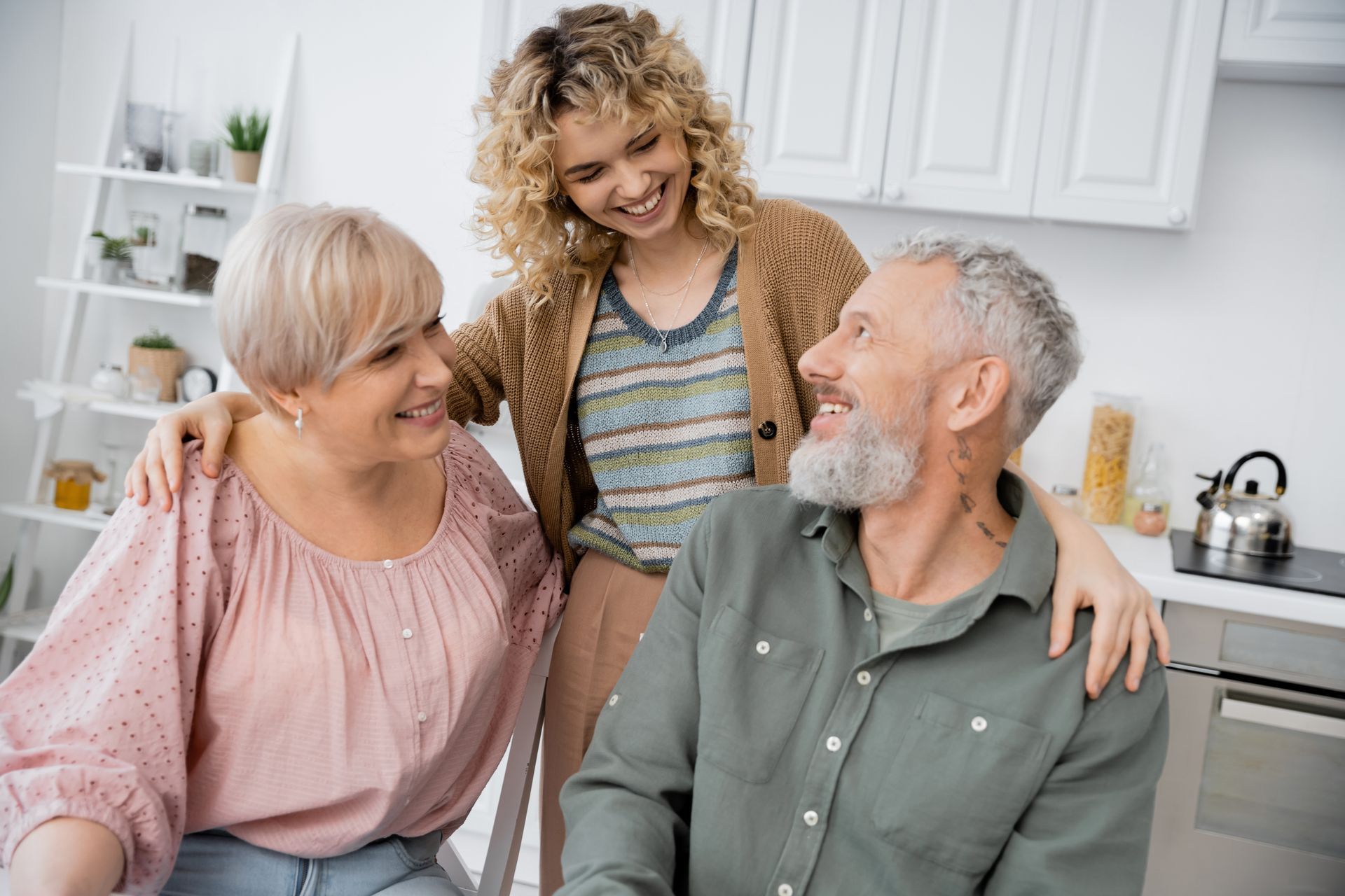 Three smiling people in a bright kitchen, with one standing behind two seated adults.