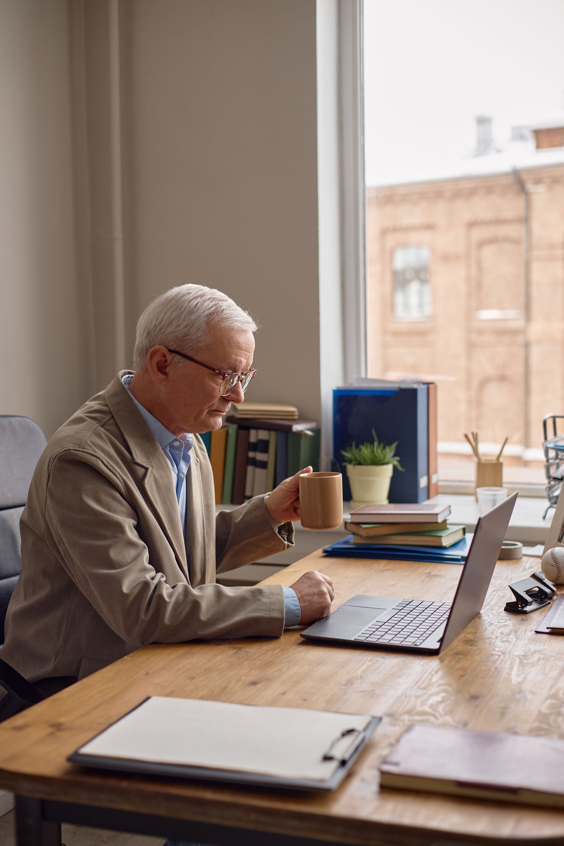 Older man working at a desk by a large window, reading papers beside a laptop and coffee mug