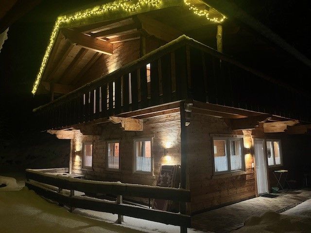 Eine zweistöckige Holzhütte bei Nacht, erleuchtet von warmem Außenlicht und Lichterketten entlang der Dachkante.