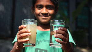 Person holding two glasses, one cloudy drink and one clear, while smiling indoors in sunlight