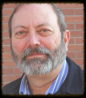 Close-up portrait of a bearded man in a blue shirt and jacket, standing before a brick wall.
