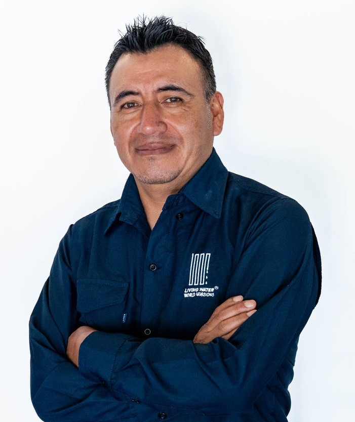 Man in blue shirt and white cap smiling indoors, holding a cloth or bag near a tiled wall