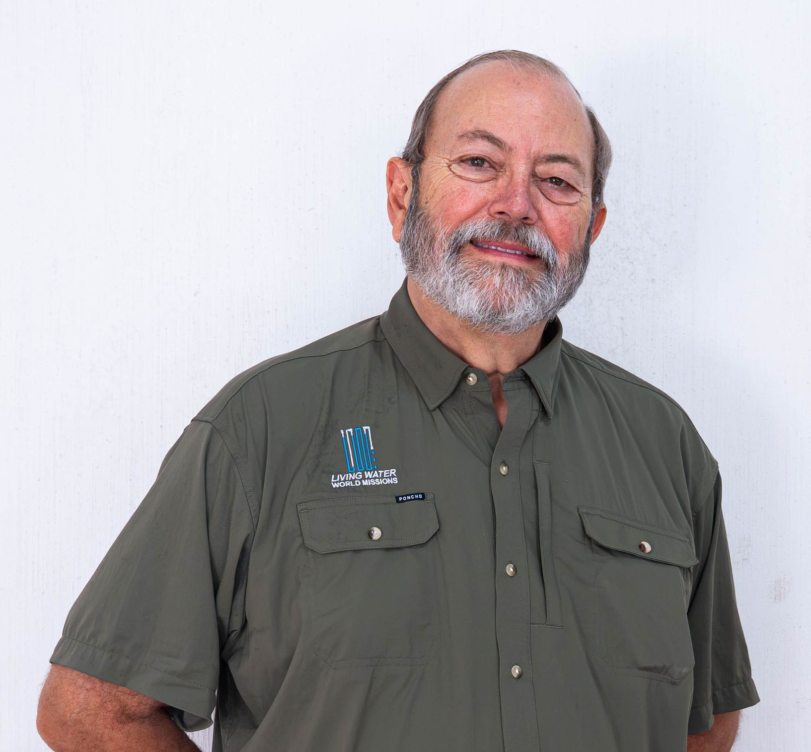 Close-up portrait of a bearded man in a blue shirt and jacket, standing before a brick wall.