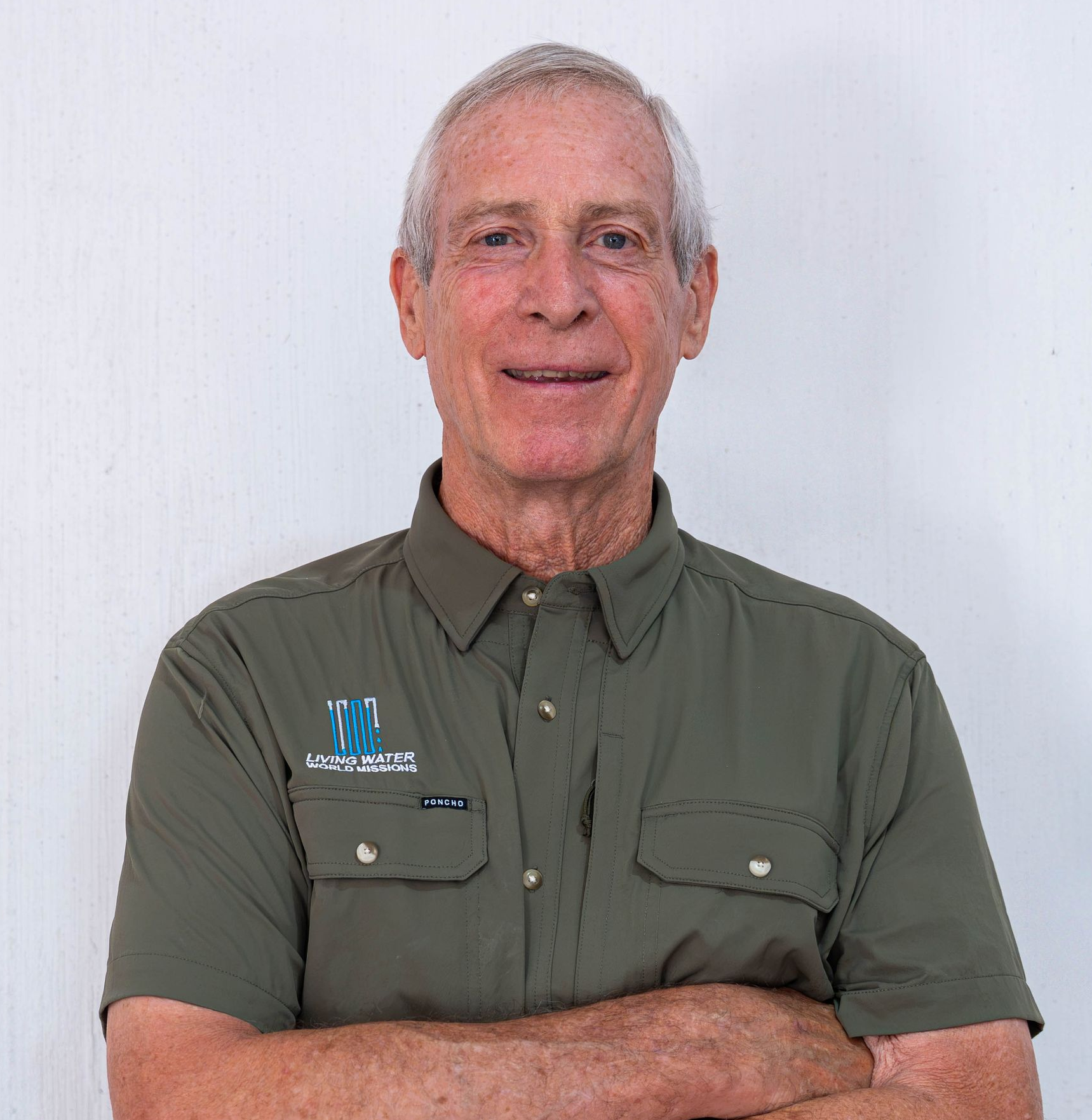 Smiling man in a light blue shirt standing outdoors with blurred green trees behind him