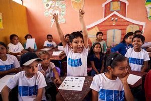 Children in a classroom raising their hands, smiling at desks with worksheets on the table