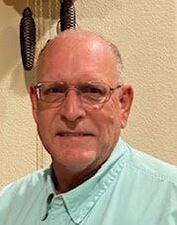 Portrait of a bald man wearing glasses and a mint-green button-up shirt against a beige wall