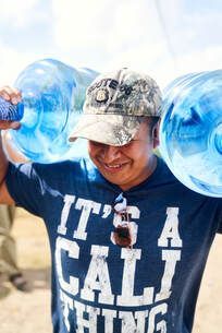 Man lifting two large water jugs on his shoulders, wearing a “IT’S A CALL” T-shirt and camouflage cap.