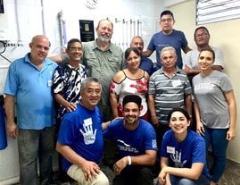 Group of nine people posing indoors, smiling and standing in two rows, some wearing blue shirts.