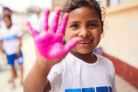 Smiling child in white shirt holds up a bright pink-painted hand indoors
