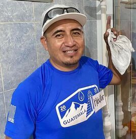 Man in blue shirt and white cap smiling indoors, holding a cloth or bag near a tiled wall