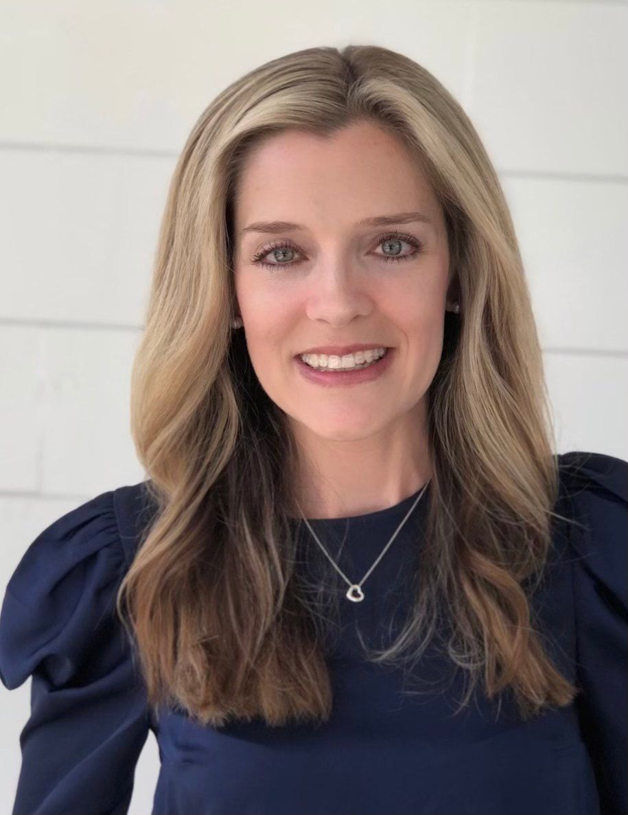 Smiling woman with long blonde hair in a navy blouse against a light wall