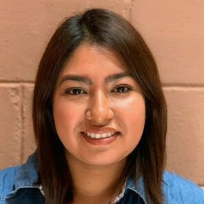 Smiling person with dark hair wearing a blue shirt against a pink brick wall