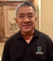 Man standing indoors in a black polo shirt, smiling at the camera.
