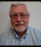 Portrait of a smiling man with white hair, glasses, and a light blue plaid shirt on a plain background