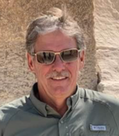 Man wearing sunglasses and a gray shirt, standing outdoors against a stone wall, smiling.