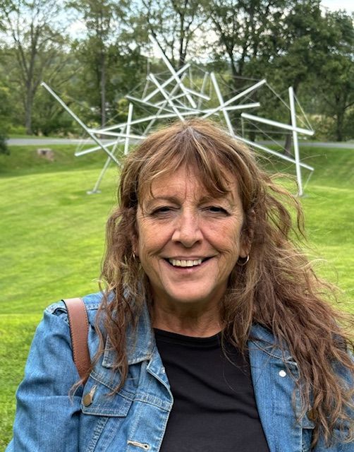 Smiling woman in a denim vest standing on a grassy lawn with a white abstract metal sculpture behind her
