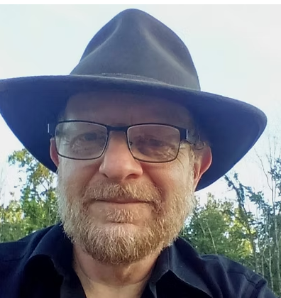 Man wearing glasses and a black hat outdoors, with trees and sky in the background