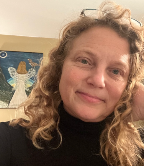 Person with curly blonde hair in a black turtleneck, smiling indoors with a colorful painting behind them