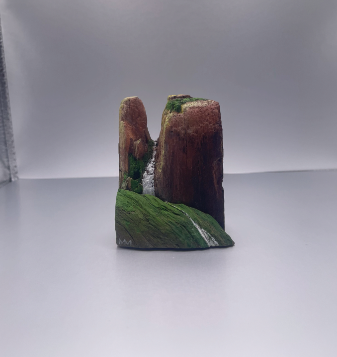 Small painted rock formation with green moss-like details on a white surface against a plain background