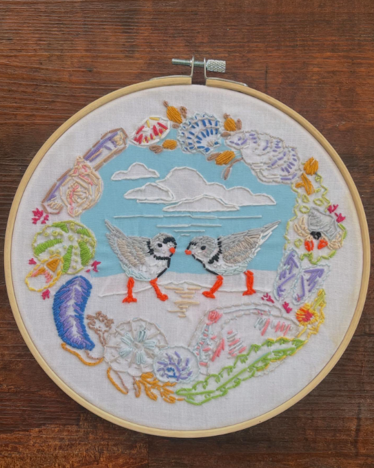 Embroidered hoop with two geese under clouds, surrounded by colorful flowers on fabric.