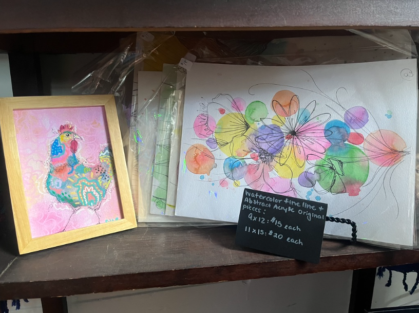 Framed peacock art and colorful floral drawing displayed on a shelf with a small chalkboard sign