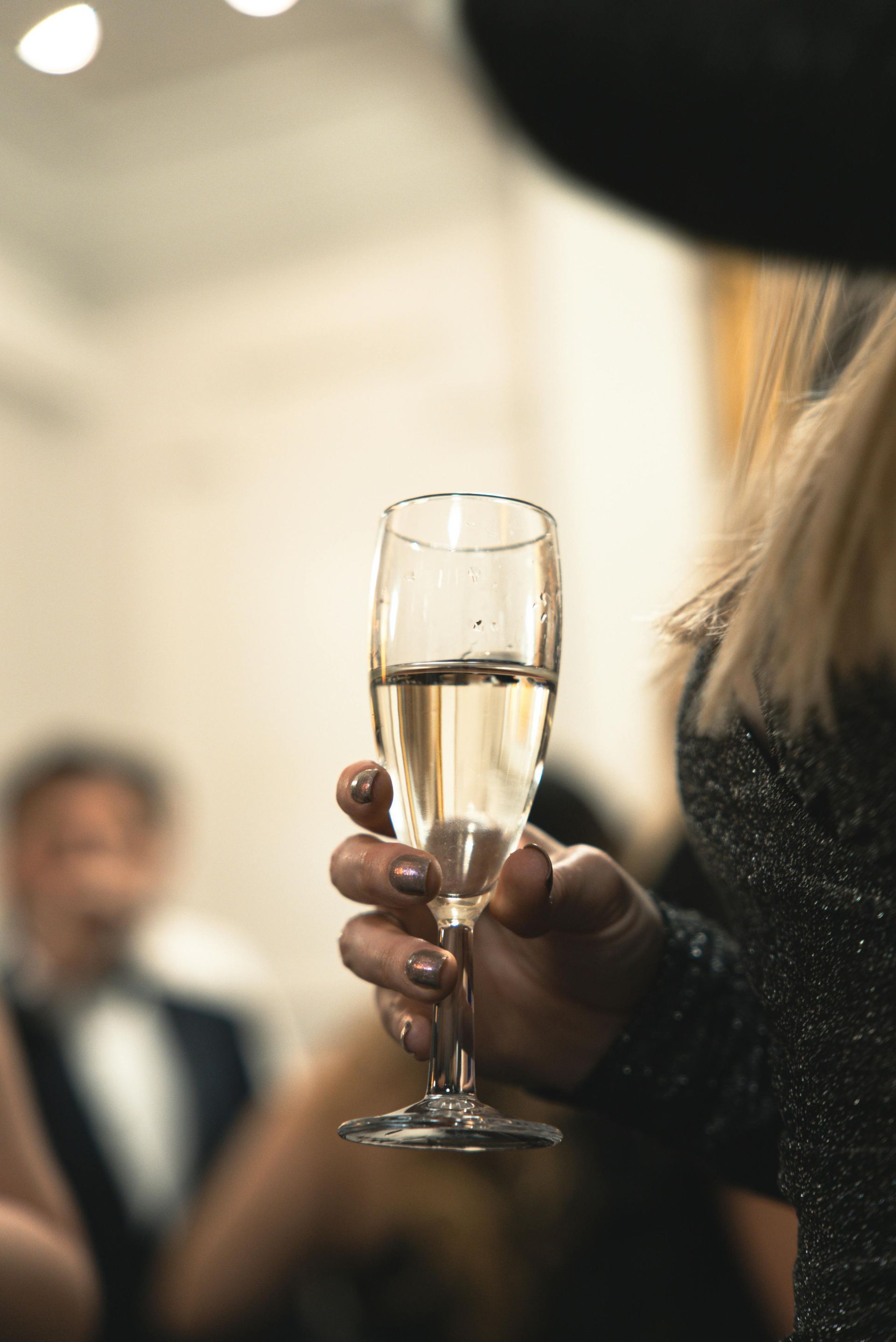 Hand holding a champagne flute at a formal event, with a blurred guest in the background