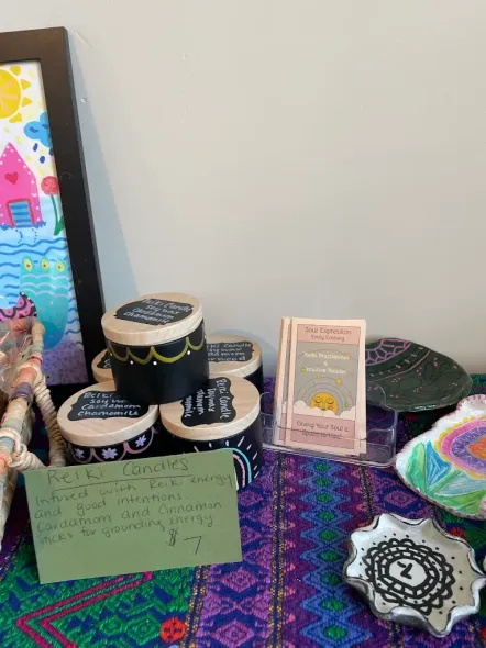Craft fair display with hand-painted black jars, colorful sign, and crochet items on a purple patterned cloth.