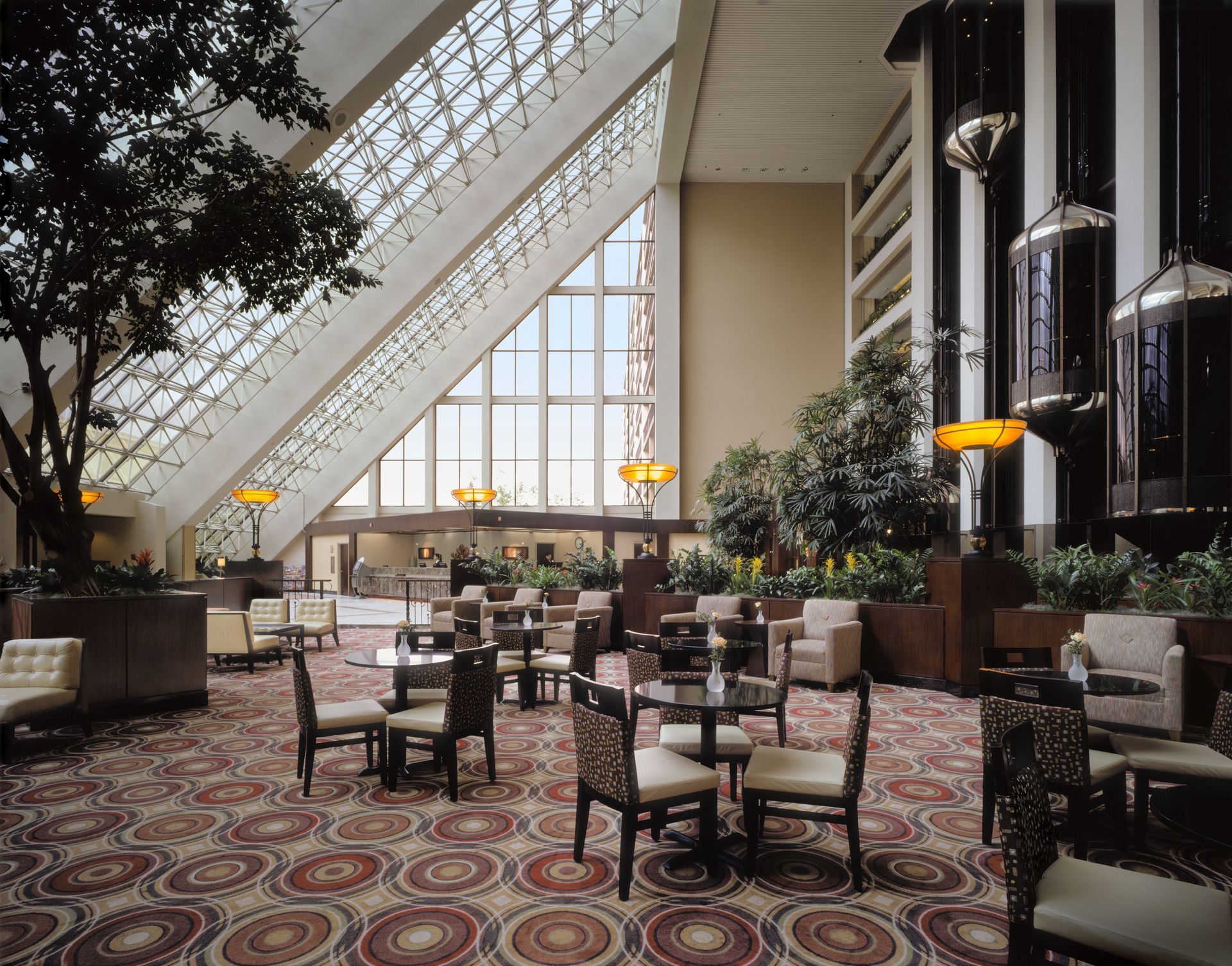 Elegant hotel atrium lounge with tables, chairs, and skylight windows under a patterned carpet.