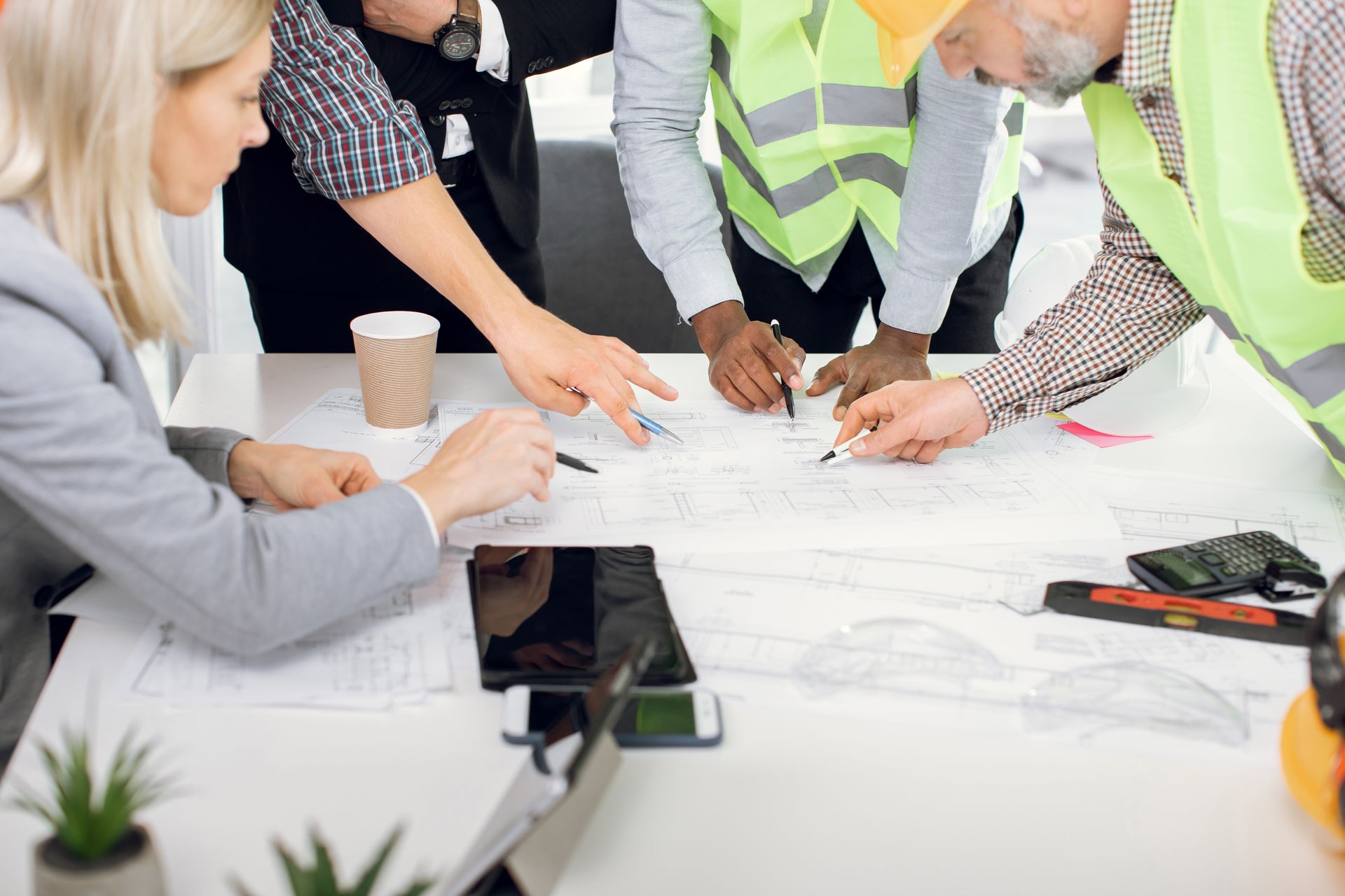 Team members in safety vests and office clothes review plans around a table with papers and a coffee cup.